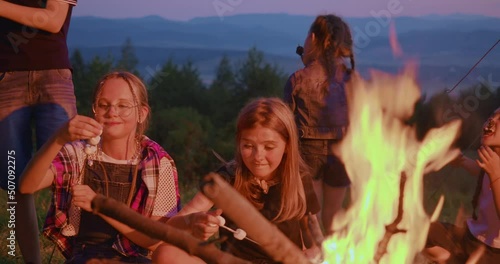 Front view of two girls sitting near campfire, stringing marshmallows on skewer for frying in mountains. Group of children eating roasted meal, having fun on summer camp at dusk. Concept of camping.