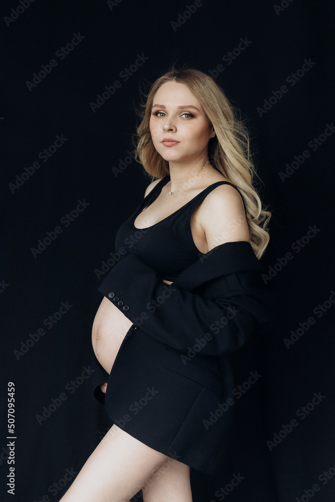a blonde pregnant woman in a black jacket and lingerie stands in the studio on a black background holds her hand on her stomach a blonde pregnant woman in a black jacket and lingerie stands in the studio on a black background holds her hand on her stomach