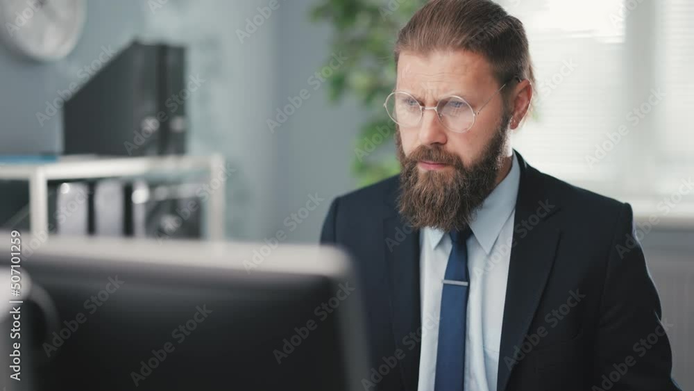 Handsome bearded man with serious face looking on monitor while typing on keyboard. Male entrepreneur in glasses and suit using modern pc for hard work at bright office center. Handsome bearded man with serious face looking on monitor while typing on keyboard. Male entrepreneur in glasses and suit using modern pc for hard work at bright office center.