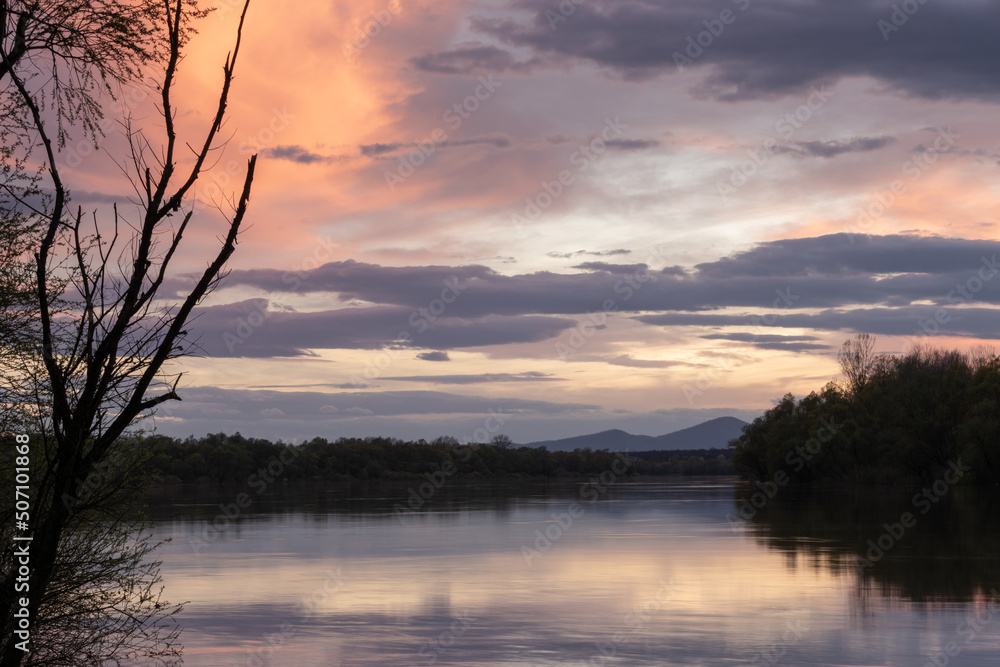 Landscape of river and mountain silhouette with cloudy sky, Sava river with forested shore and Motajica mountain during dusk with orange glow in clouds