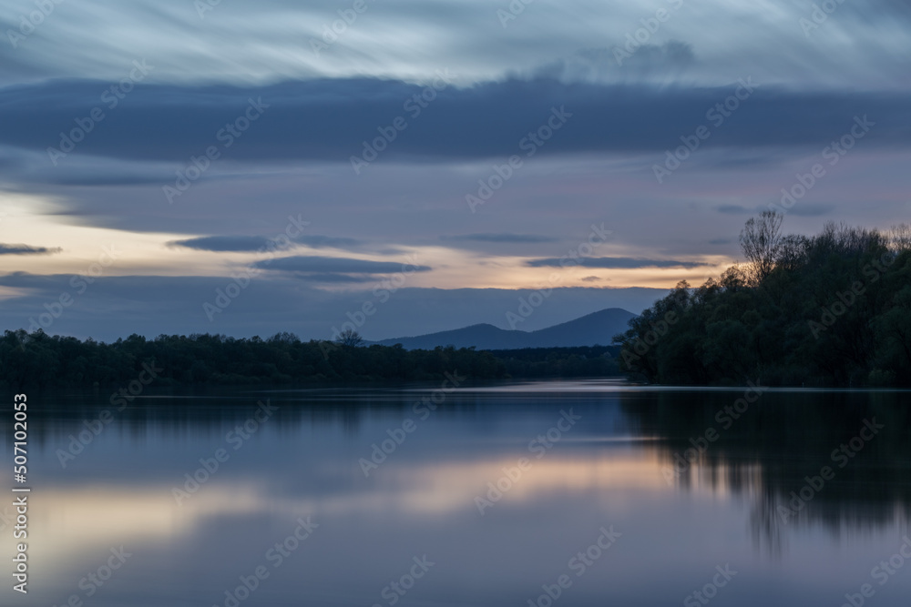 Landscape of river and mountain silhouette at twilight, Sava river with forested shore and Motajica mountain scene with clouds in sky during blue hour in long exposure