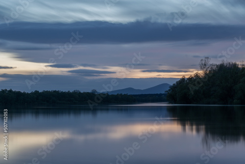 Landscape of river and mountain silhouette at twilight, Sava river with forested shore and Motajica mountain scene with clouds in sky during blue hour in long exposure