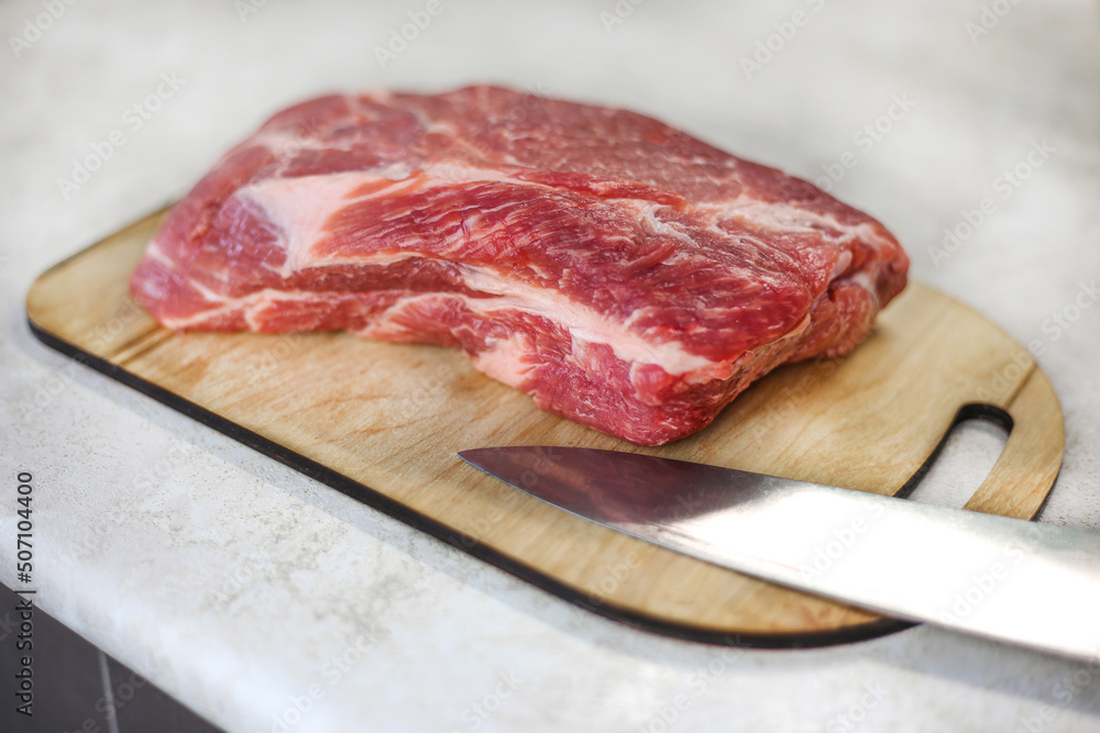 Large fresh piece of meat on a wooden cutting board with a metal knife on a light kitchen table Large fresh piece of meat on a wooden cutting board with a metal knife on a light kitchen table