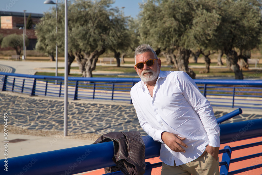 Mature, gray-haired man with beard, sunglasses, white shirt and leather jacket, leaning on a railing outdoors. Concept grandfather, sugar daddy, lifestyle, mature man. Mature, gray-haired man with beard, sunglasses, white shirt and leather jacket, leaning on a railing outdoors. Concept grandfather, sugar daddy, lifestyle, mature man.