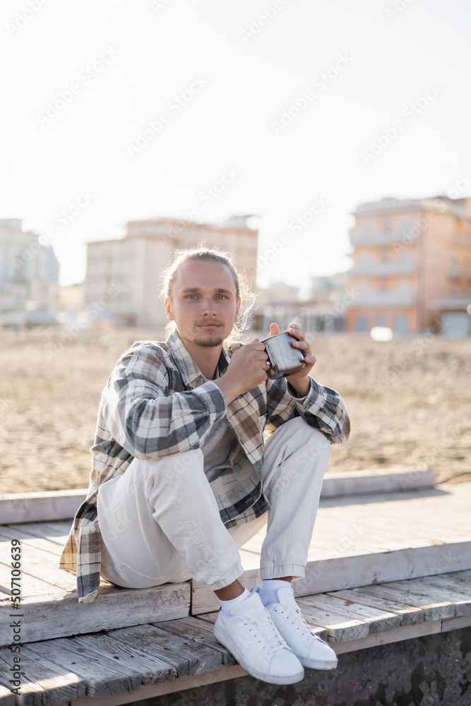 Young man in shirt holding metal cup while sitting on pier in Treviso.