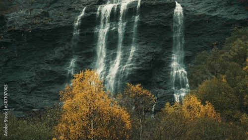 Thin fog hangs above the waterfall on the Aselvi river. Whitewater cascading from dark cliffs. Colorful autumn trees below. Slow-motion, pan left. photo