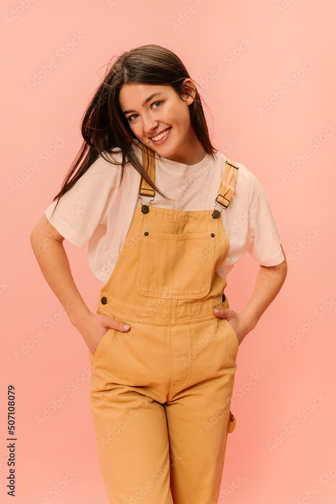 Cute young caucasian girl is smiling looking into camera with hands in pockets on pink background. Brunette with loose hair is wearing white T-shirt and yellow jumpsuit. People's emotions concept. Cute young caucasian girl is smiling looking into camera with hands in pockets on pink background. Brunette with loose hair is wearing white T-shirt and yellow jumpsuit. People's emotions concept.