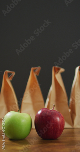 Vertical image of red and green apple with row of packed lunches in paper bags and grey background