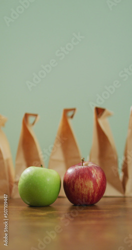 Vertical image of red and green apple with row of packed lunches in paper bags and green background