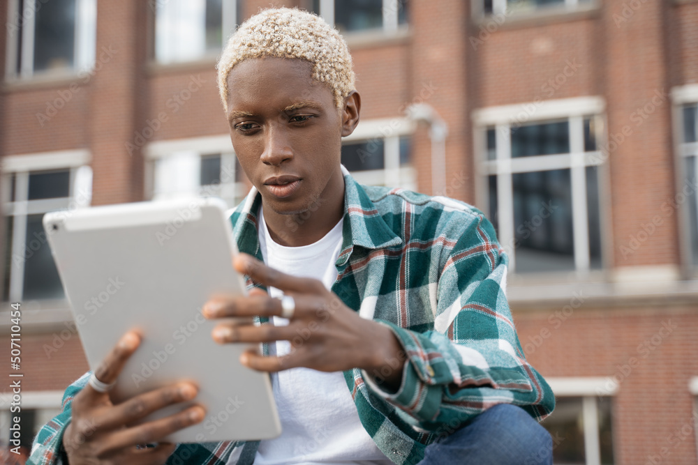 Portrait of young pensive African American man with stylish hair using digital tablet watching videos sitting on the street. Student studying, online education concept Portrait of young pensive African American man with stylish hair using digital tablet watching videos sitting on the street. Student studying, online education concept