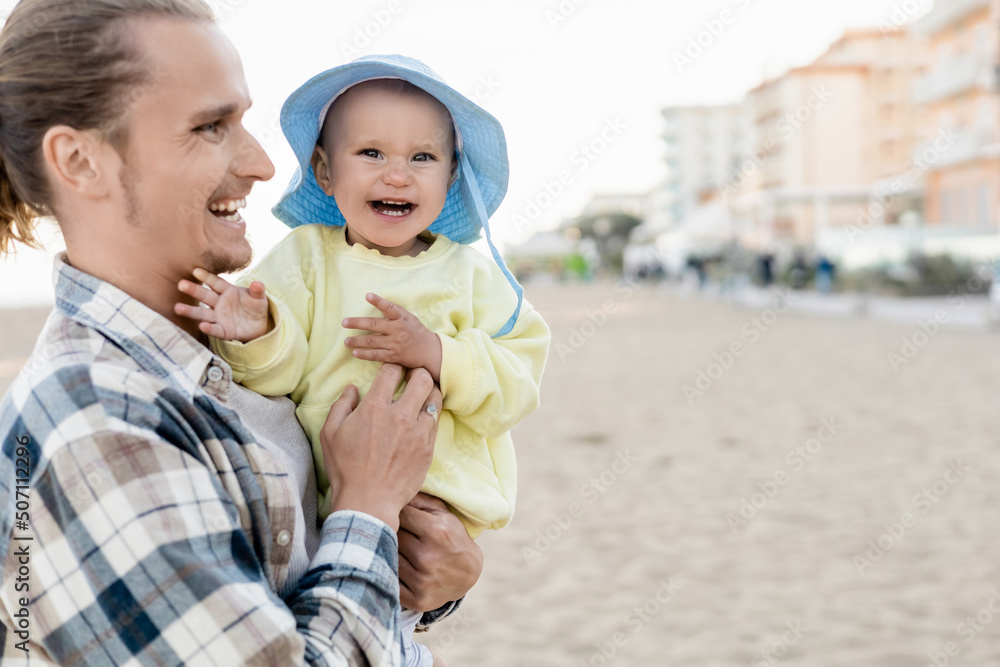 Fototapeta premium Positive man holding baby girl in panama hat on beach in Treviso.