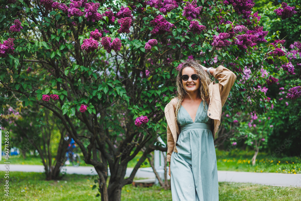 A beautiful curly woman in a summer dress runs and rejoices, she is wearing sunglasses. Against the background of a purple lilac bush A beautiful curly woman in a summer dress runs and rejoices, she is wearing sunglasses. Against the background of a purple lilac bush