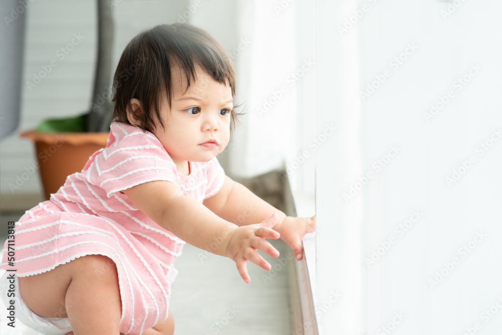 Adorable little mixed race, Caucasian and Asian, toddler baby girl wear pink dress and diaper is sitting on floor try to standing and looking out of window with curious face Adorable little mixed race, Caucasian and Asian, toddler baby girl wear pink dress and diaper is sitting on floor try to standing and looking out of window with curious face