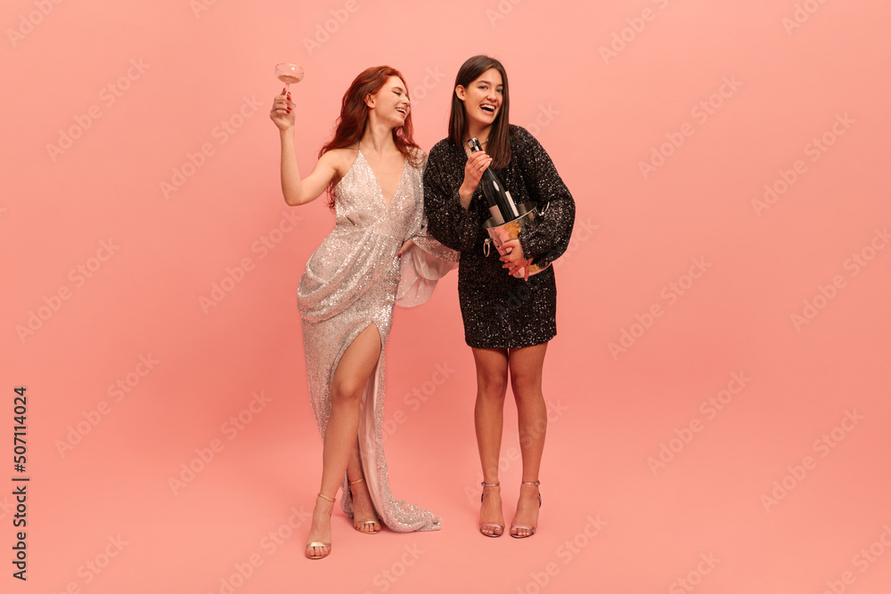 Full-length young caucasian redheaded lady raises her glass, brunette holding champagne against pink background. Models wearing evening gowns celebrate prom. Concept of holiday Full-length young caucasian redheaded lady raises her glass, brunette holding champagne against pink background. Models wearing evening gowns celebrate prom. Concept of holiday