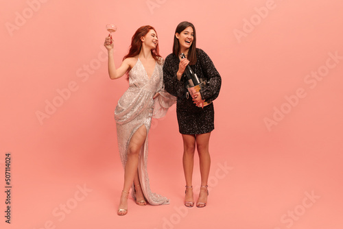 Full-length young caucasian redheaded lady raises her glass, brunette holding champagne against pink background. Models wearing evening gowns celebrate prom. Concept of holiday