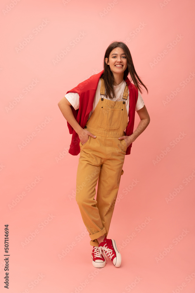 Full-length modern young caucasian girl smiles looking at camera against pink background. Brunette girl wears T-shirt, jumpsuit and sweatshirt over her shoulders. Lifestyle, leisure concept. Full-length modern young caucasian girl smiles looking at camera against pink background. Brunette girl wears T-shirt, jumpsuit and sweatshirt over her shoulders. Lifestyle, leisure concept.