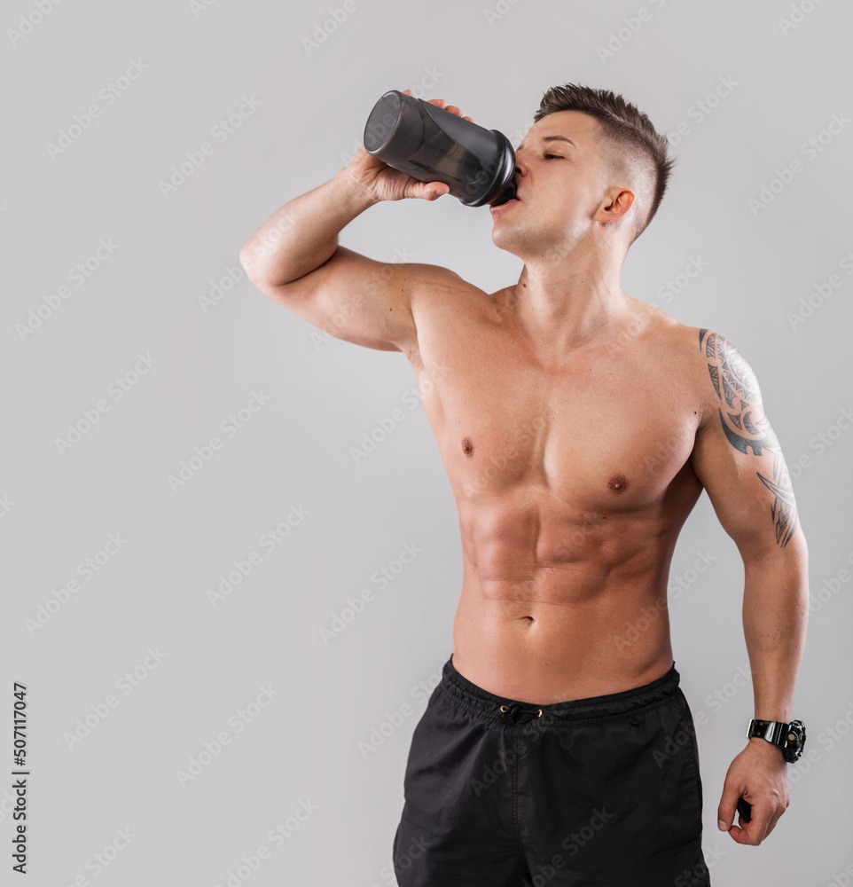 Naklejka premium Athletic muscular handsome guy with a naked sporty body with a tattoo drinking water from a black bottle against a gray background. Healthy lifestyle and diet