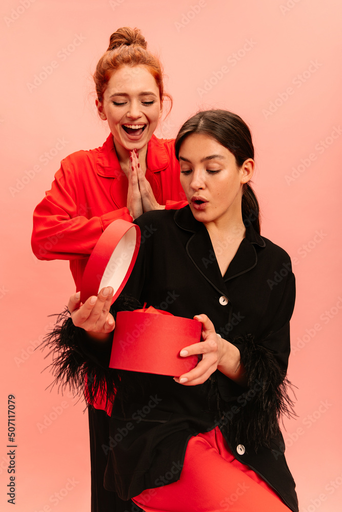 Two young caucasian girls with positive emotions look at gift on pink background. Brunette looks into box in surprise, redhead happily holds her palms together. Two young caucasian girls with positive emotions look at gift on pink background. Brunette looks into box in surprise, redhead happily holds her palms together.