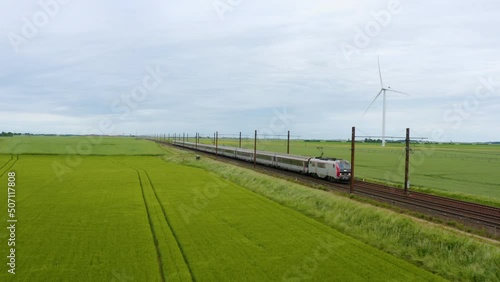 Aerial view of local train traveling through green fields under sunset sky and giant wind turbines. Train, railway in the meadow, windmills in the distance, sunset sky, Drone 4K. High-speed train. 