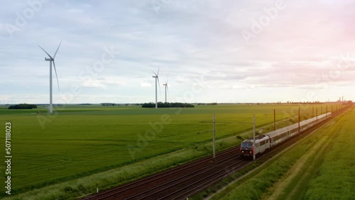 Aerial view of local train traveling through green fields under sunset sky and giant wind turbines. Train, railway in the meadow, windmills in the distance, sunset sky, Drone 4K. High-speed train. 
