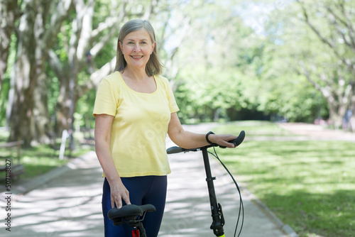 Portrait of senior active woman, gray-haired woman in park looking at camera and smiling with bicycle, summer day