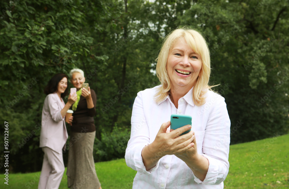Old blond lady watches the news on her phone, in the distance in the background her female friends also use smartphones. Old blond lady watches the news on her phone, in the distance in the background her female friends also use smartphones.