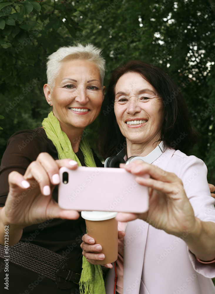 Two charming senior women friends holds a cup of coffee to go and making selfie in summer park. Two charming senior women friends holds a cup of coffee to go and making selfie in summer park.