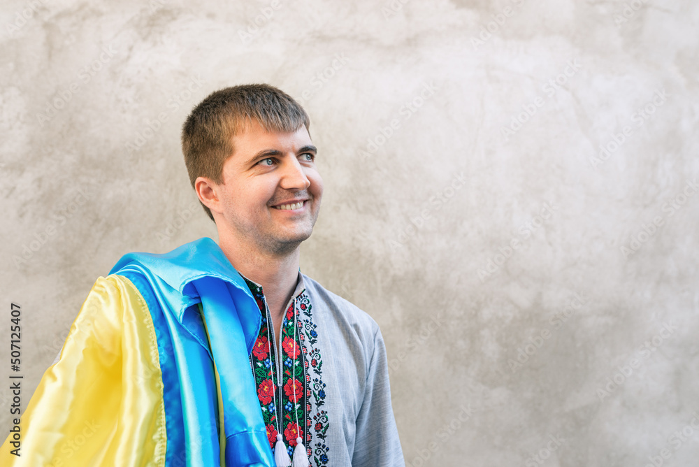 Portrait of a smiling man holding the blue and yellow State Flag of Ukraine on his shoulder. Guy in a traditional embroidered shirt on a background concrete wall. Day of the Constitution. Copy space. Portrait of a smiling man holding the blue and yellow State Flag of Ukraine on his shoulder. Guy in a traditional embroidered shirt on a background concrete wall. Day of the Constitution. Copy space.