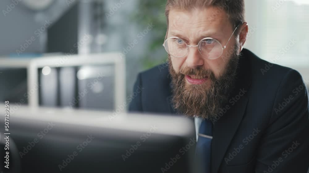 Close up of focused bearded man wearing glasses and business suit sitting at office desk and having conference call. Caucasian male employer talking with his workers using modern pc. Close up of focused bearded man wearing glasses and business suit sitting at office desk and having conference call. Caucasian male employer talking with his workers using modern pc.