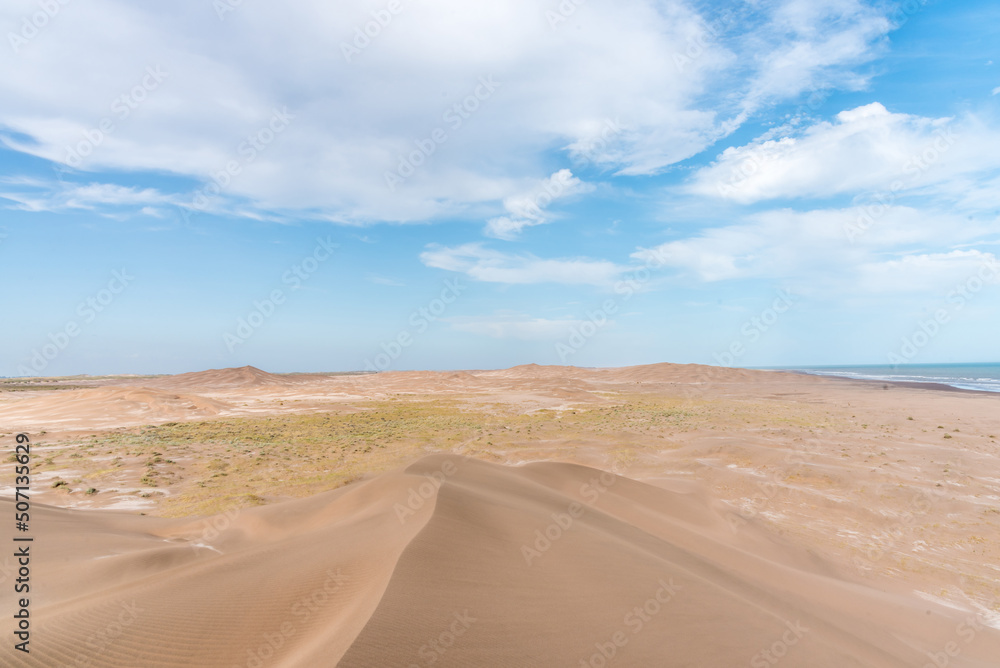 Dunes in front of the beach with the blue sky in the background and the horizon in the sea with some clouds.