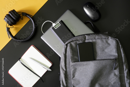 Compact urban backpack with laptop, external battery - power bank, mobile phone, headphones, mouse, notepad and pen on black and yellow background. Study, travel and trip with gadgets.