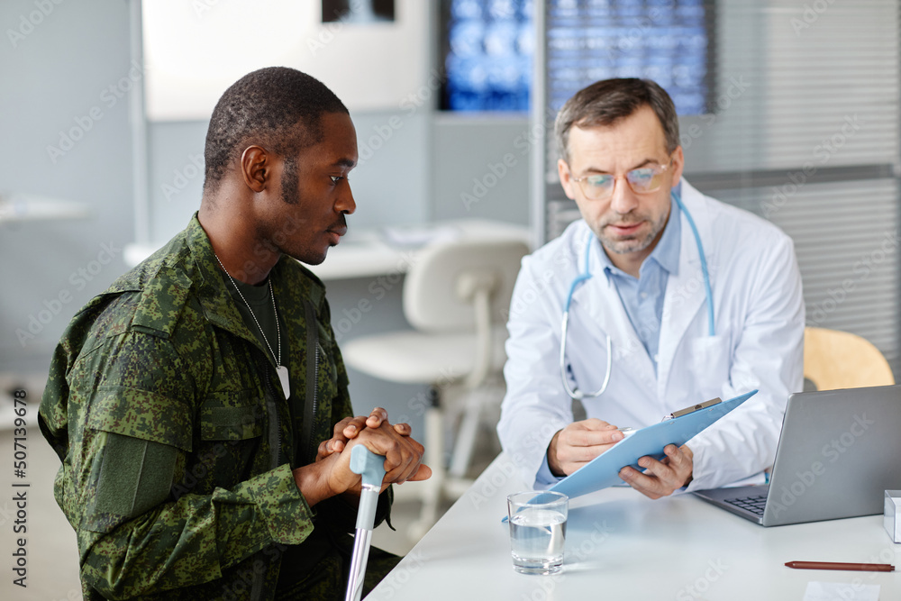 Mature doctor explaining checkup test results to young Black military man using walking stick after getting injured Mature doctor explaining checkup test results to young Black military man using walking stick after getting injured