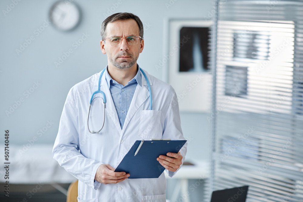 Horizontal medium portrait of serious mature Caucasian medical specialist wearing lab coat and stethoscope holding clipboard looking at camera Horizontal medium portrait of serious mature Caucasian medical specialist wearing lab coat and stethoscope holding clipboard looking at camera