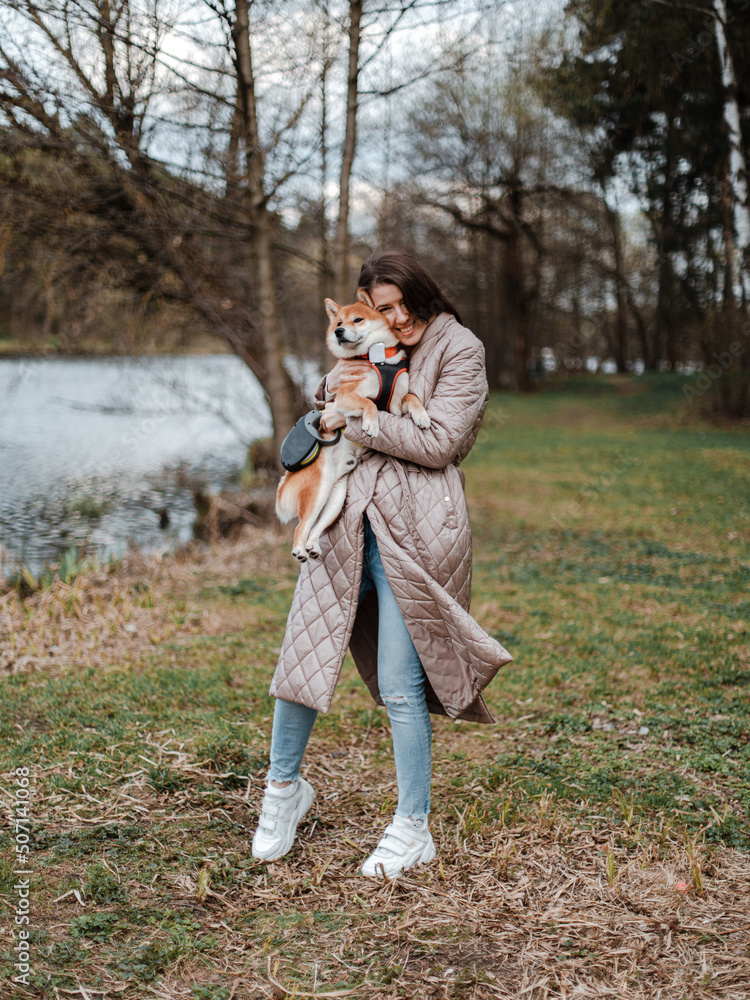 Fototapeta premium Young millennial beautiful woman brunette is walking with her Japanese breed shiba inu dog outdoor near the riverside bank on a leash. 