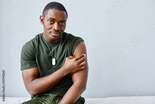 Portrait of strong and healthy young African American soldier sitting on medical couch pressing piece of cotton pad on shoulder after getting vaccine injection