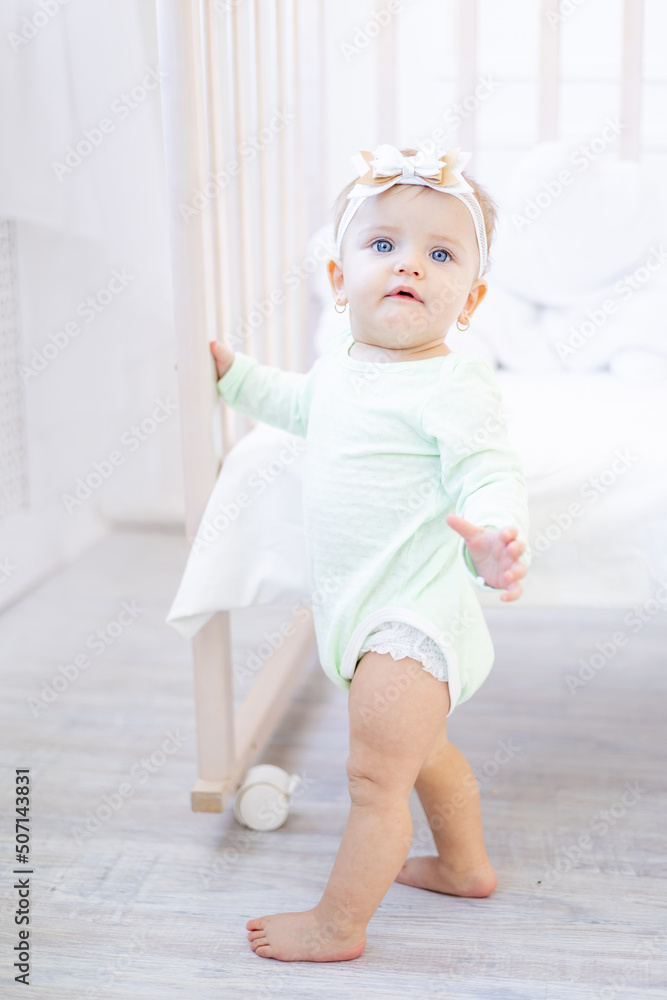 cute little baby girl is standing at the crib in the nursery in a green cotton bodysuit and learning to walk taking her first steps