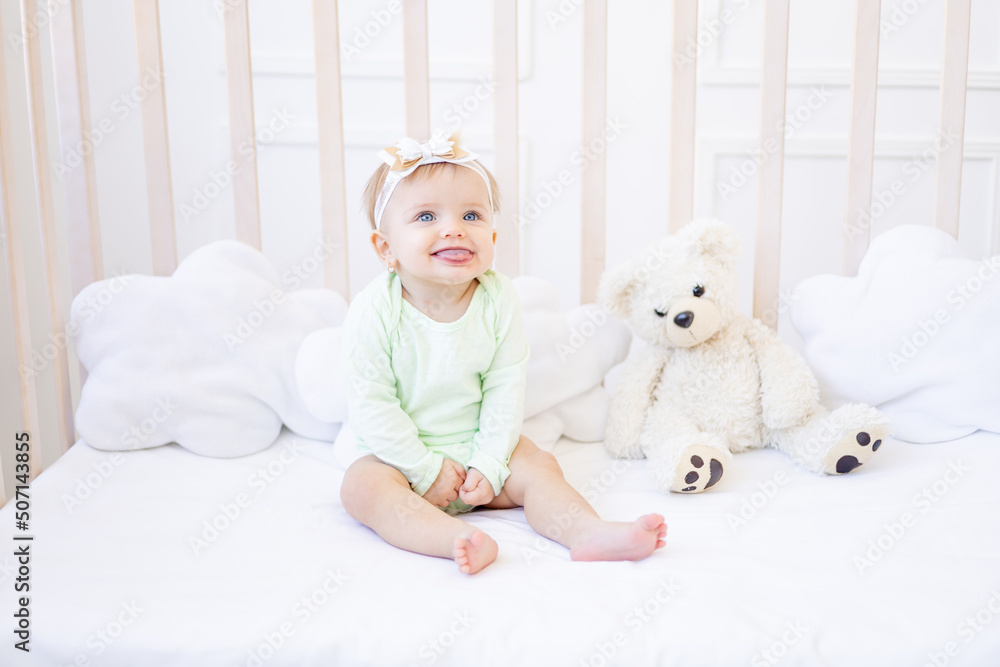 cute funny little baby girl shows tongue in a crib on a white cotton bed in a green bodysuit in a children's bright room and smiles cute funny little baby girl shows tongue in a crib on a white cotton bed in a green bodysuit in a children's bright room and smiles