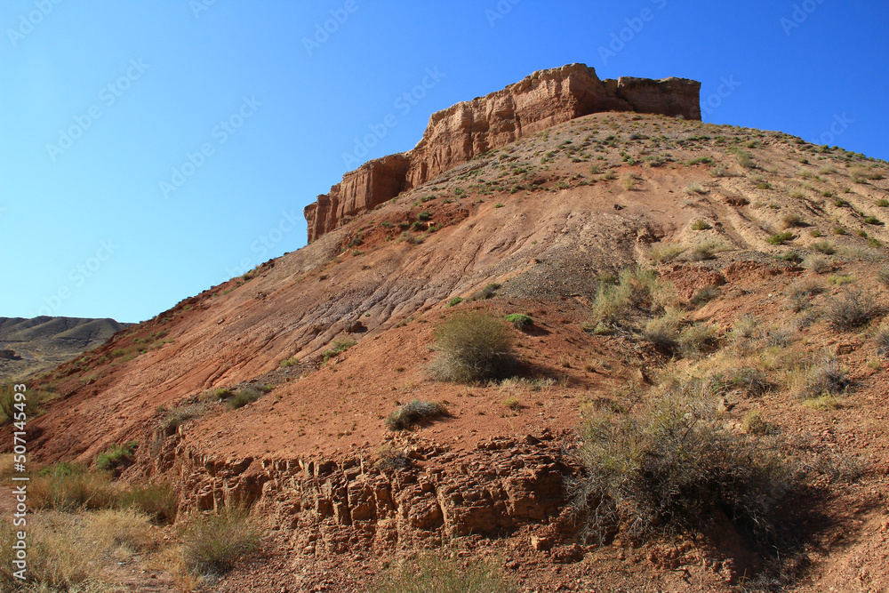 In the Temirlik canyon there is a large red sandy-clay mountain, on top of a rock in the form of walls and towers, bushes on the slopes, summer, sunny