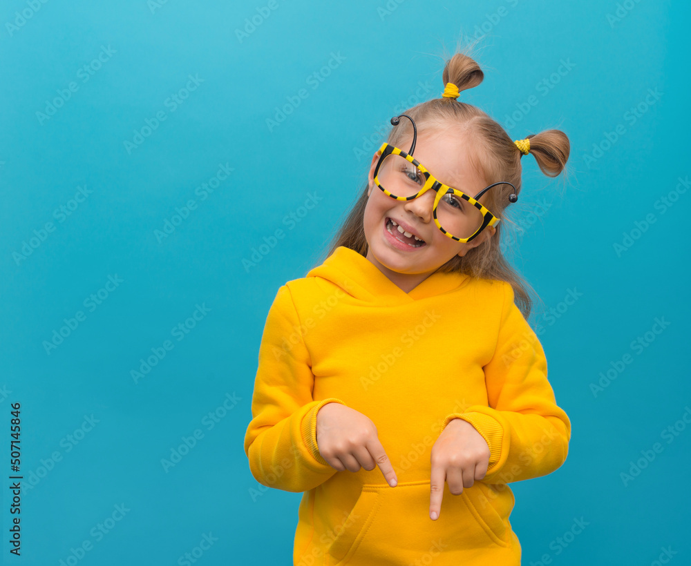 Naklejka premium Funny playfull little girl in bee glasses and yellow hoodie on blue background pointing down to free copy space.
