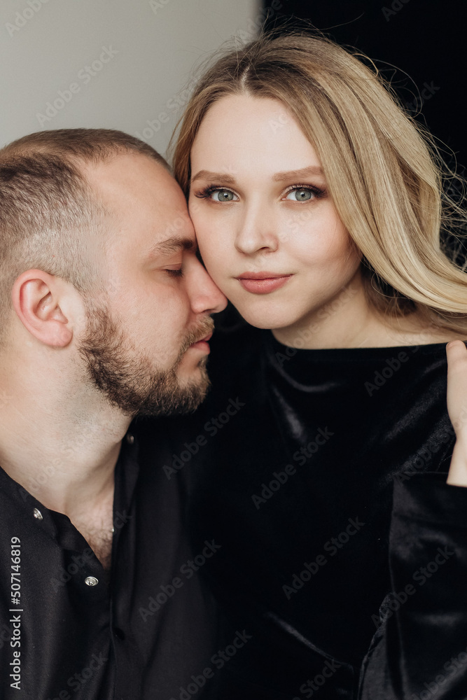 Husband and pregnant blonde wife in a black dress in the studio on a black background sit on a chair and hug Husband and pregnant blonde wife in a black dress in the studio on a black background sit on a chair and hug