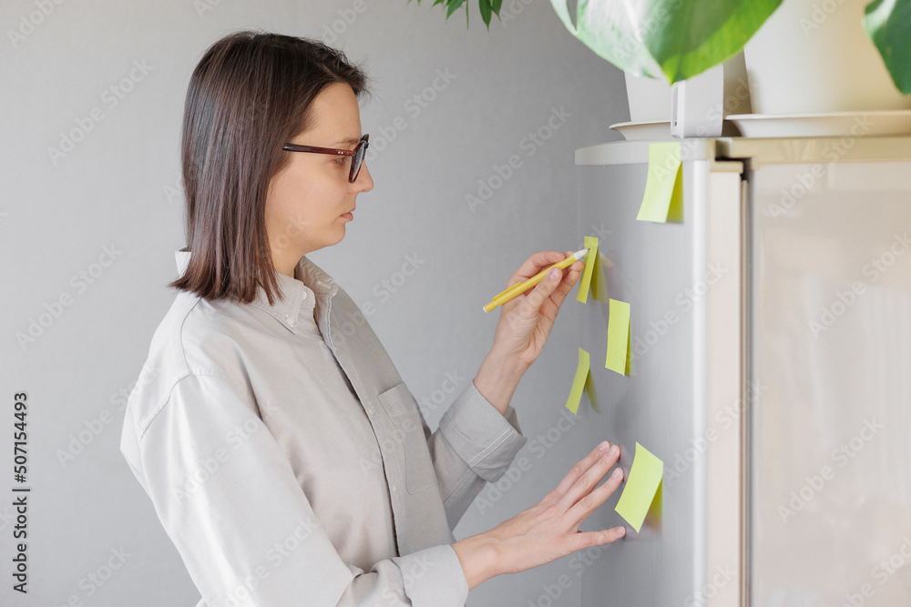 a woman makes notes on colored stickers on the refrigerator. a reminder or a note for her husband and children. home planning of tasks and a list of products to buy in the store. homemaker management a woman makes notes on colored stickers on the refrigerator. a reminder or a note for her husband and children. home planning of tasks and a list of products to buy in the store. homemaker management