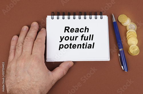 On a brown surface are coins, a pen and a notepad with the inscription - reach your full potential