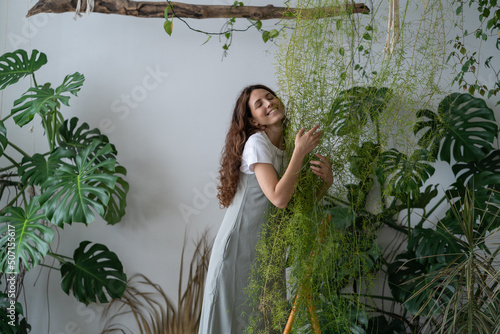 Obraz na plátně Young Spanish girl stands among green plants on stepladder and hugs flower in living room