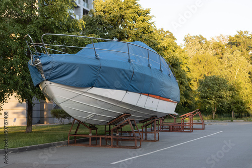 A motor boat covered with a blue cover stands on land under the open sky against the background of trees