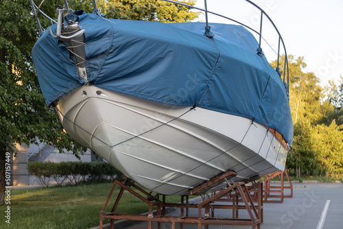 A motor boat covered with a blue cover stands on land under the open sky against the background of trees