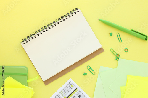 An open notepad with blank white sheets next to yellow sticky notes in a green pen and a green notepad on a bright yellow background. top view, flat lay