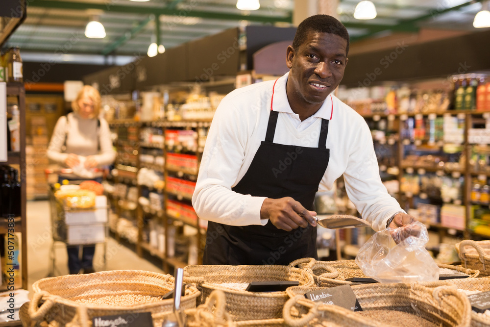 Friendly African man working in bulk food department of supermarket..