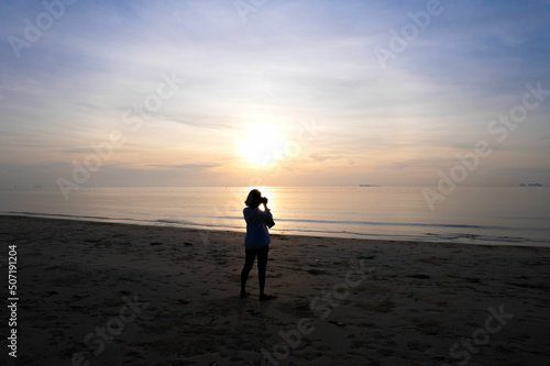 silhouette of young woman tourist she is taking pictures sun sets on the seaside The sun sets on the ground Touching the sea surface is very impressive and dazzling with paradise beach, Andaman Sea.
