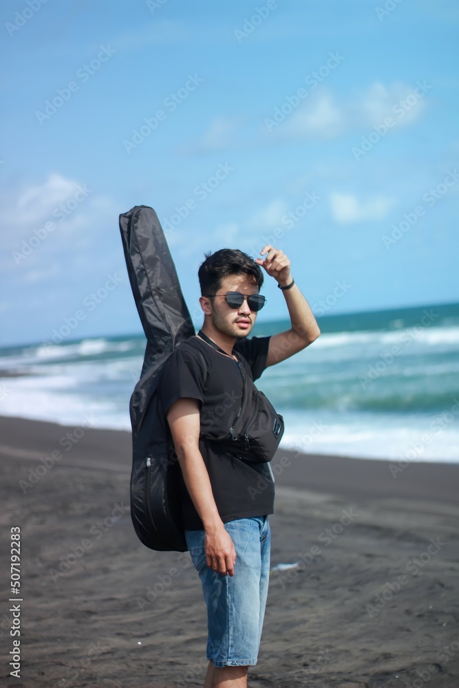 Handsome man wearing sunglasses holding guitar bag on beach