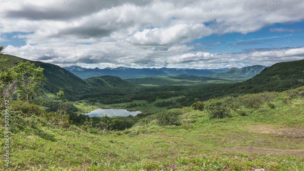 View of the valley and lake from the top of the mountain. The path goes downhill. Green vegetation around. Picturesque clouds in the blue sky. Kamchatka. Vachkazhets. Lake Tahkoloch